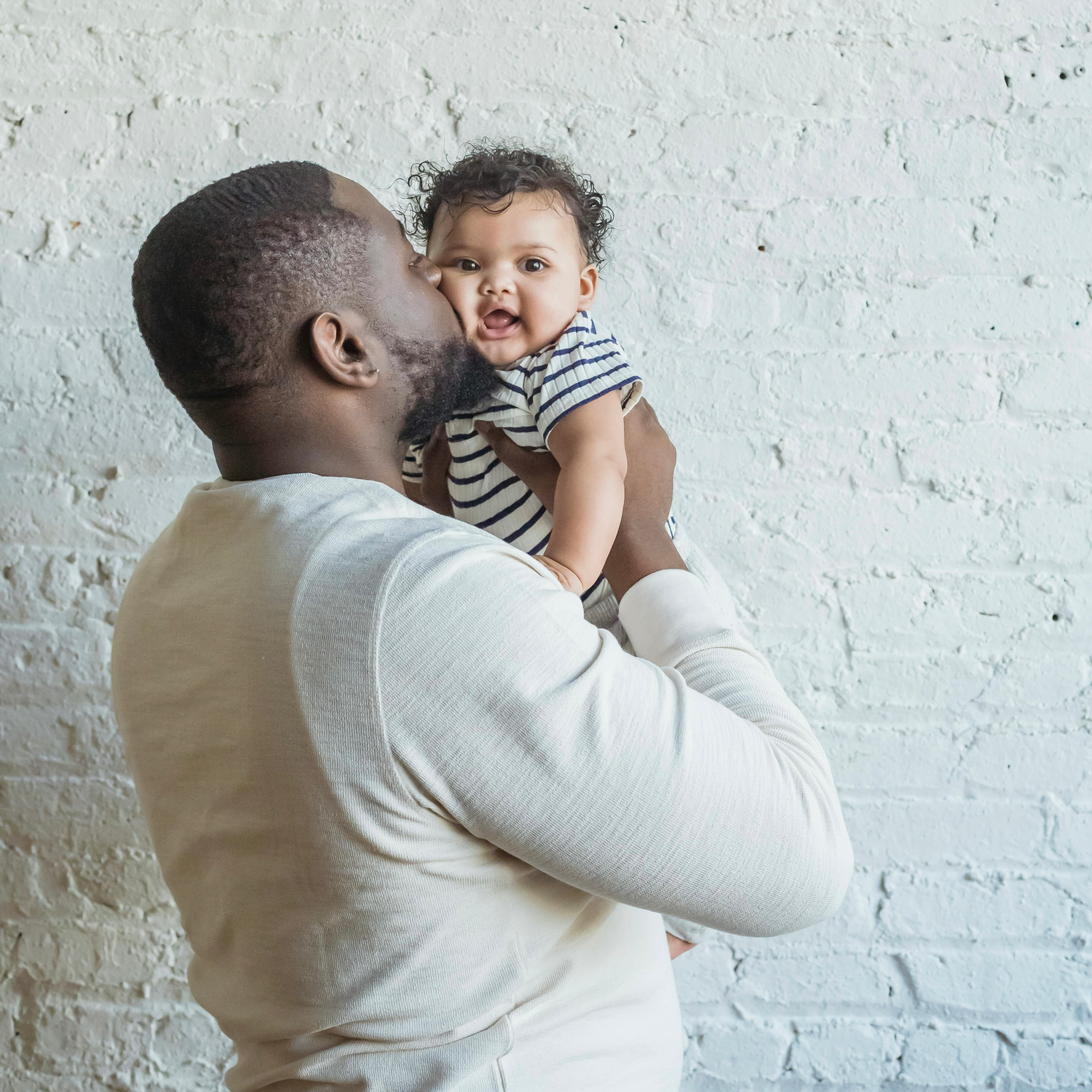 Glücklicher Vater hebt sein lachendes Baby hoch und küsst es sanft vor heller Backsteinwand – innige Familien­momentaufnahme, Kinderwunsch.