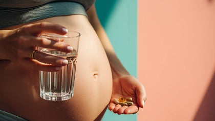 Schwangere hält ein Glas Wasser und Kapseln vor dem Babybauch vor pastelligem Hintergrund, Symbolbild für Wonder Produktqualität und Nährstoffe in der Schwangerschaft.