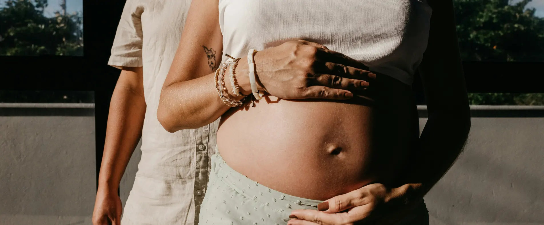 Hände liegen schützend auf einem schwangeren Bauch, daneben steht der Partner im Hintergrund bei warmem Sonnenlicht, Bild für Wonder Schwangerschaft Nährstoffe.