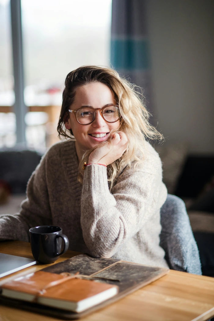 Lächelnde Content Creatorin mit Brille sitzt im Homeoffice am Schreibtisch mit Laptop und Kaffee.