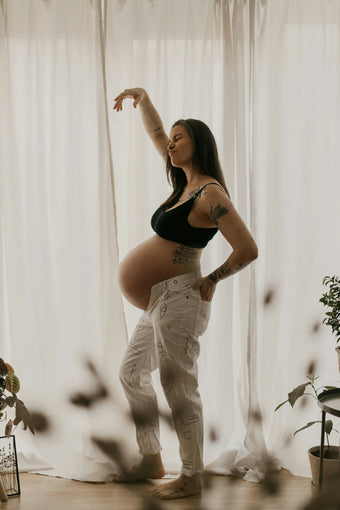 Schwangere Frau mit Tattoos steht tanzend vor hellen Vorhängen, schwarzes Top und weiße Hose, Symbolbild für Wonder Schwangerschaft Nährstoffe.