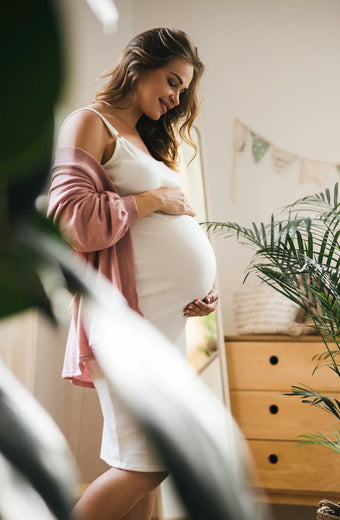 Schwangere Frau in weißem Kleid und rosa Cardigan hält ihren Babybauch zu Hause, Symbolbild für Wonder Schwangerschaft Nährstoffe.