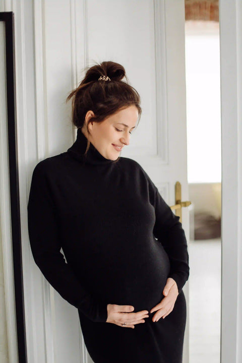 Schwangere Kundin in schwarzem Rollkragen hält ihren Bauch und lächelt, Kundenfoto einer positiven Rezension auf der Wonder Seite Produktqualität.